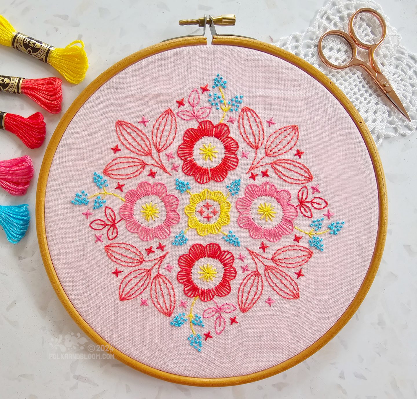 Overhead view of an embroidery in red, pink, turquoise and yellow colours on a pink fabric background.