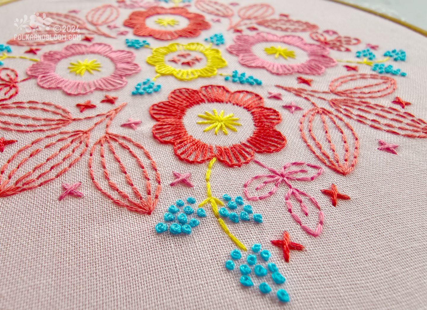 Very close view of an embroidery in red, pink, turquoise and yellow colours on a pink fabric background.