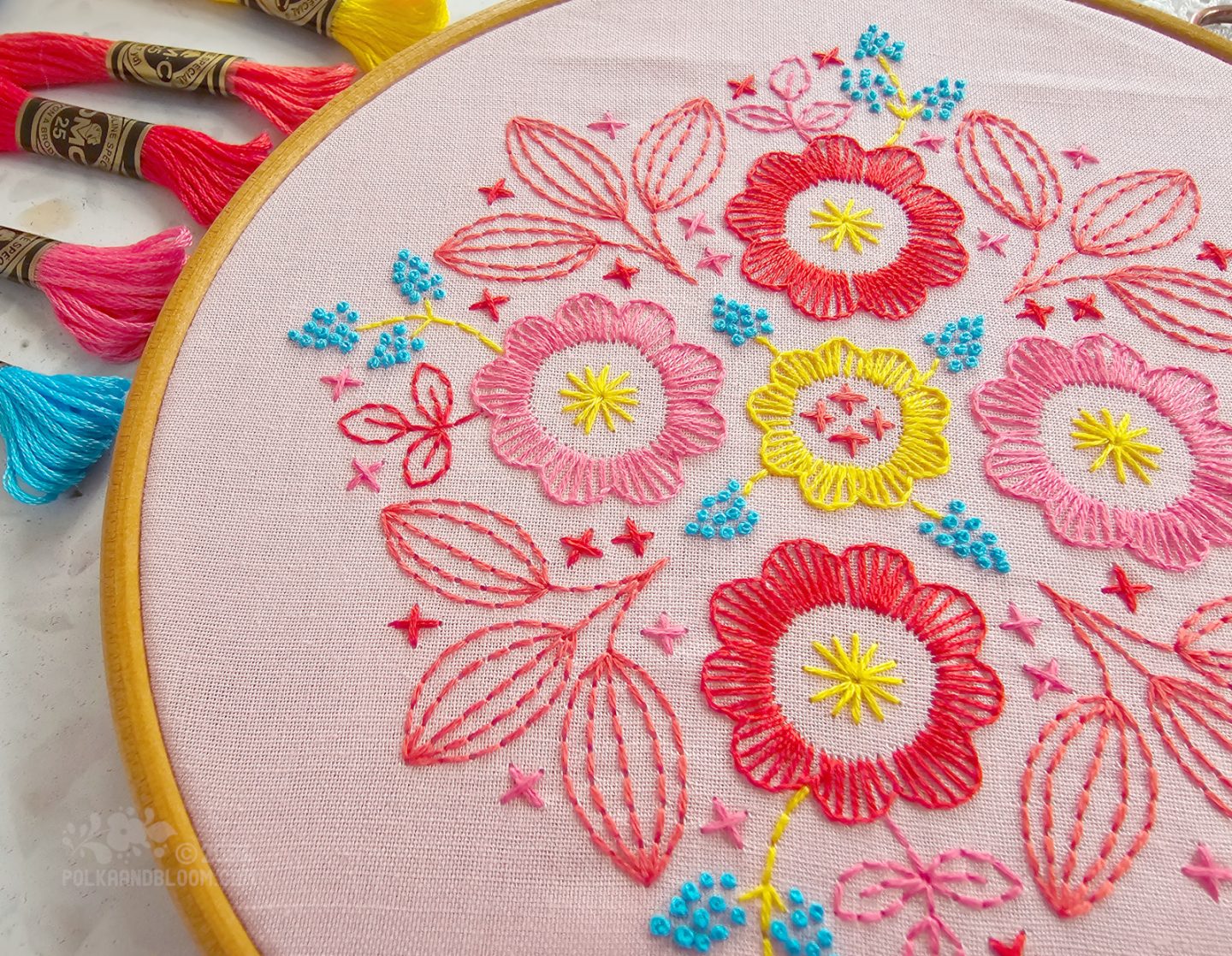 Close view of an embroidery in red, pink, turquoise and yellow colours on a pink fabric background.