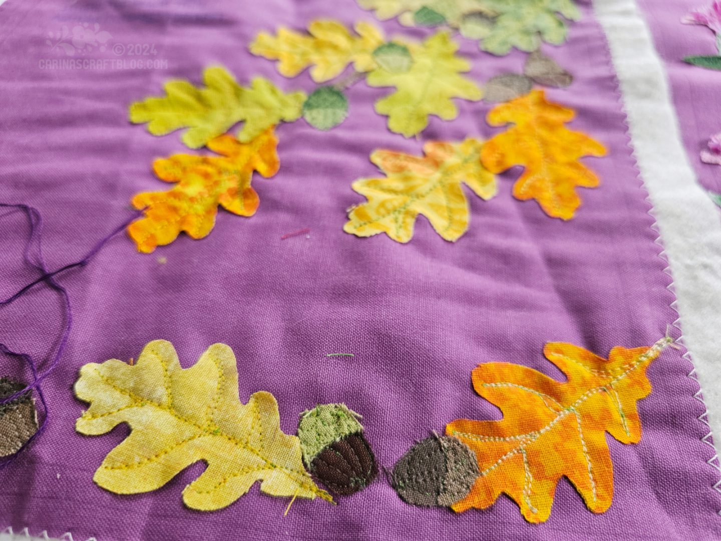 Close view of a purple square of fabric with oak leaves in orange, yellow and green appliquéd on it.