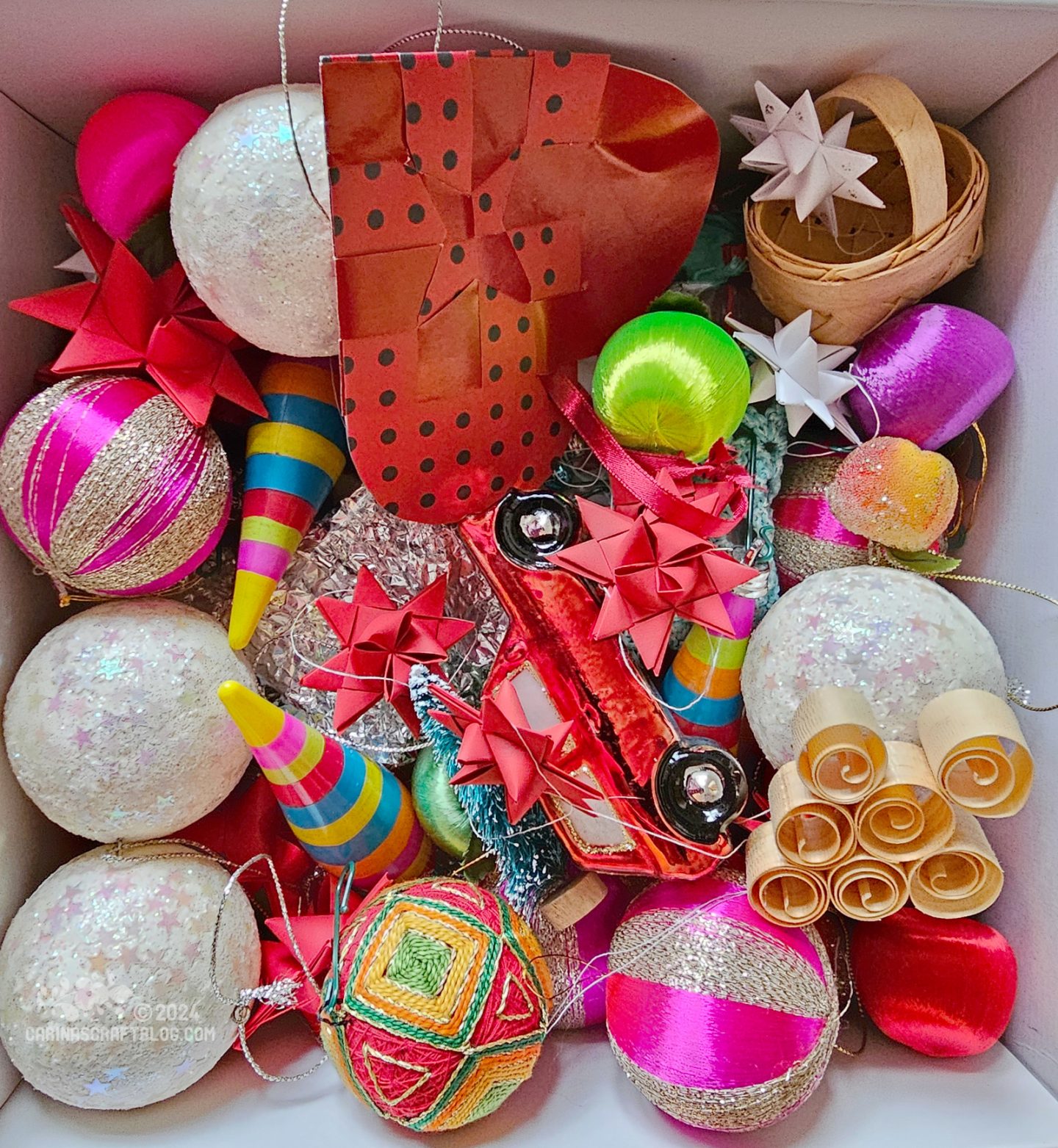 Close up photo of a box with brightly coloured Christmas ornaments: paper stars, baubles, wood shaving ornaments and a small car with a Christmas tree on its roof.