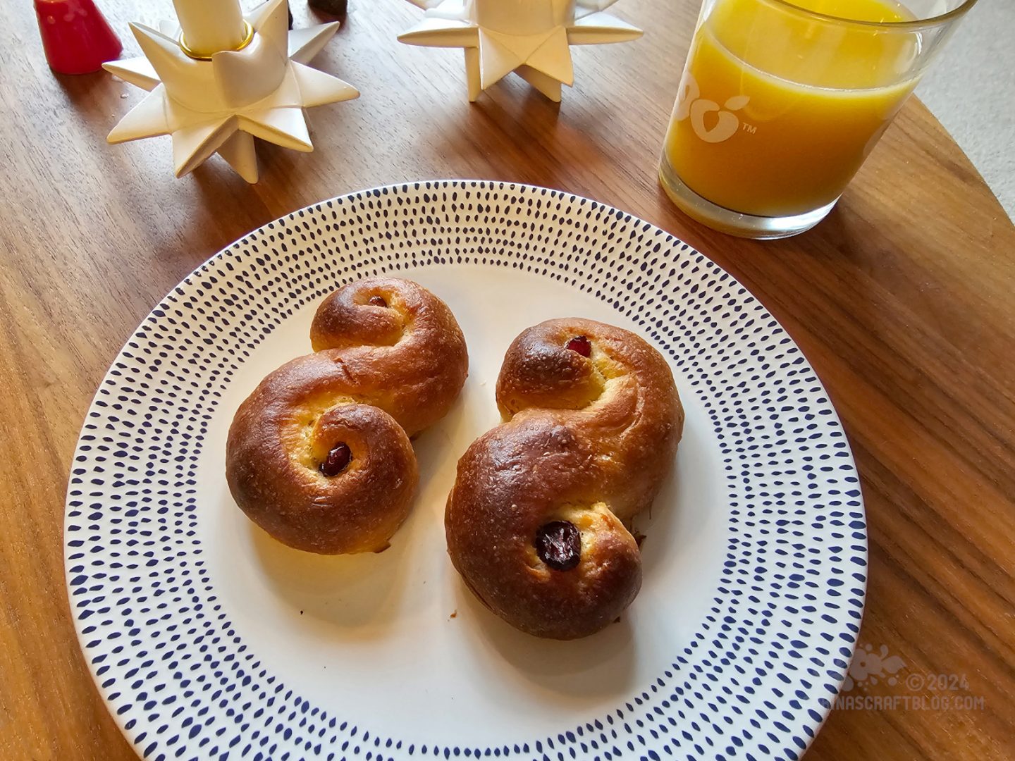 Close side view of a white plate with blue dots around the edges. On the plate are two lussekatter, saffron buns, in the shape of an S.