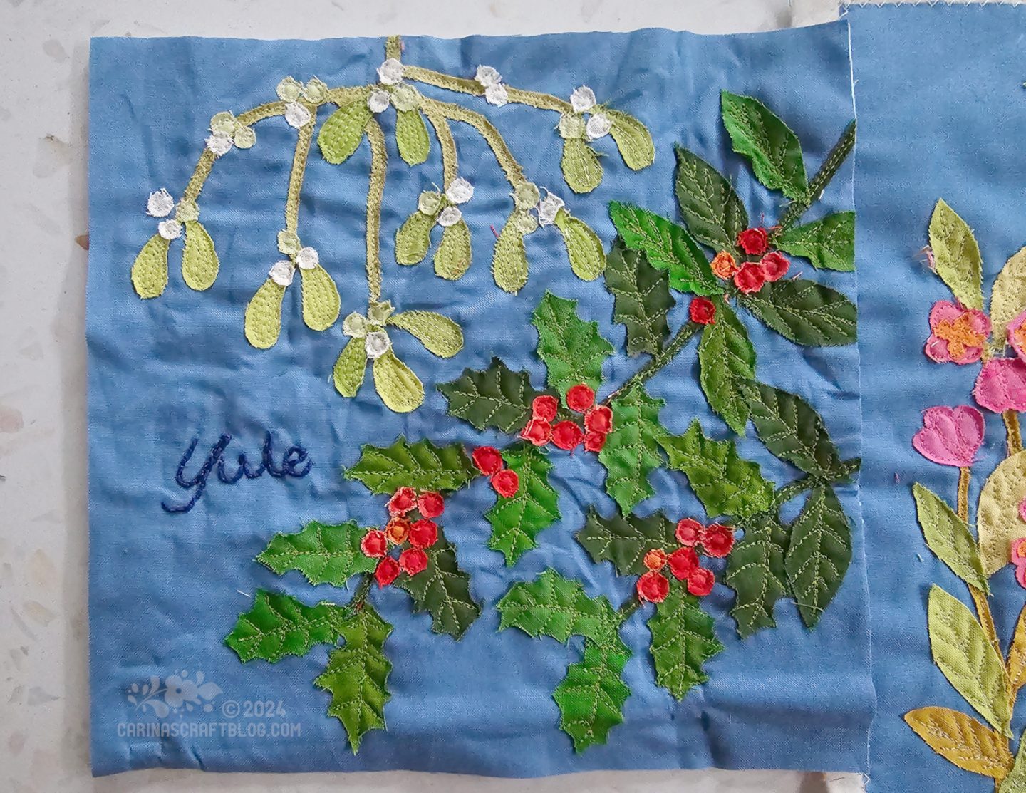 Close overhead view of a medium blue fabric square with raw edge appliqué of holly in the bottom right half and mistletoe in the top left half of the photo.