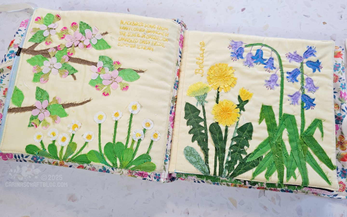 Overhead view of an open page spread of a textile book. The pages are light yellow with various flowers appliquéd on them.