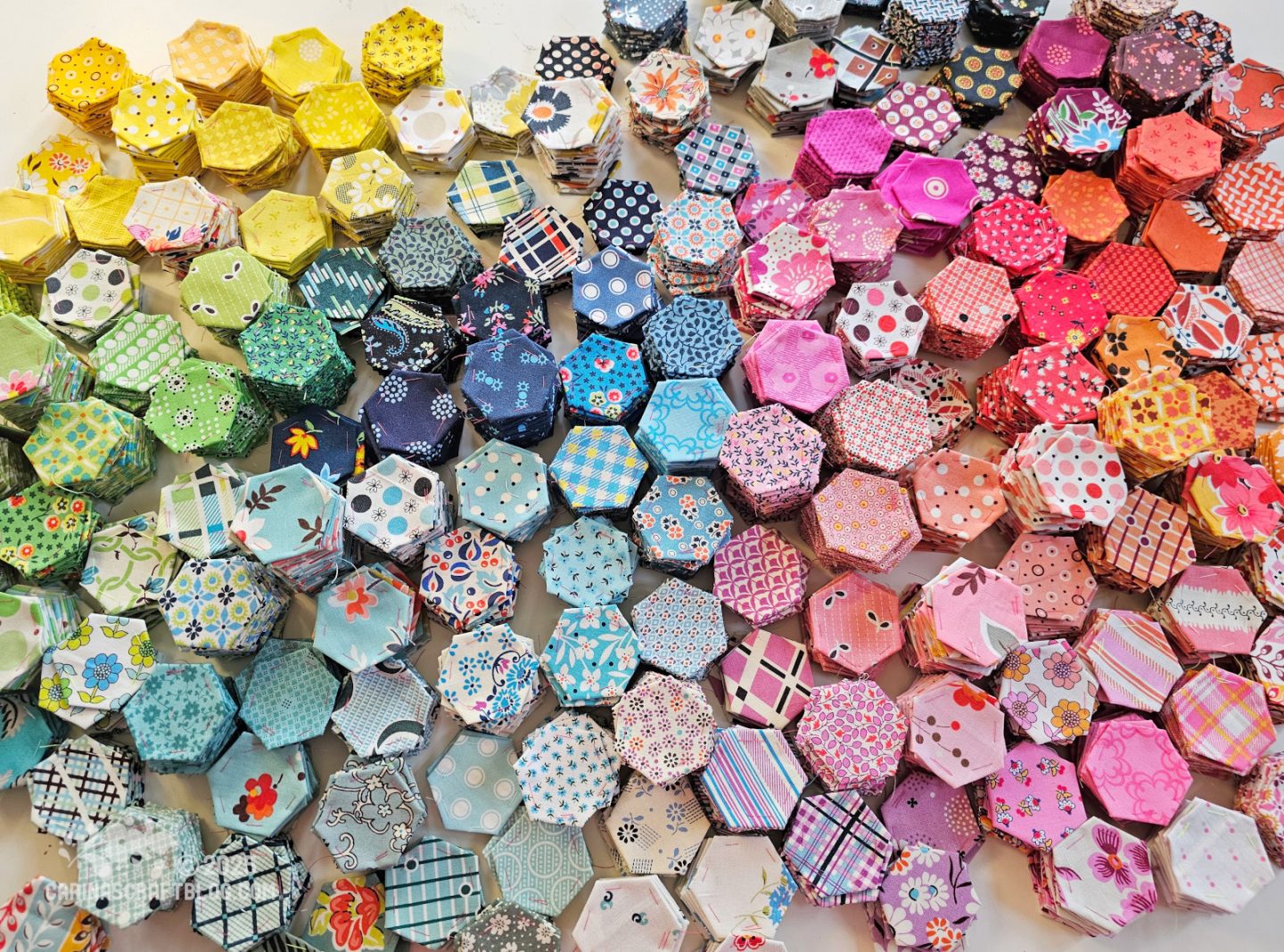 Overhead view of a table with more than a hundred stacks of fabric hexagons sorted by colour.