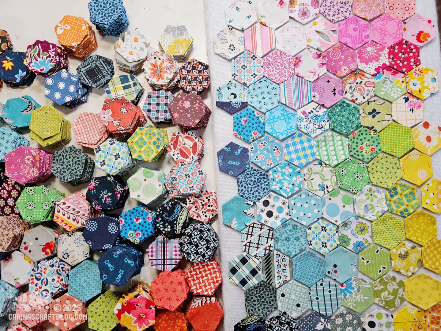 Photo. Overhead view of a white table with many stacks of fabric hexagons in many different colours, on the left hand side. On the right hand side are individual hexagons laid out side by side in a colourwash pattern.