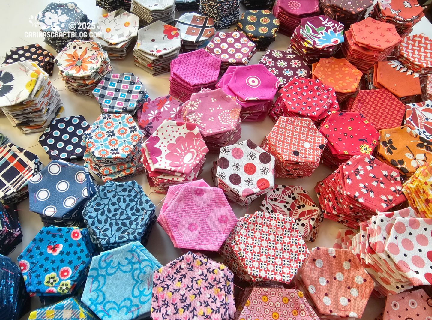 Photo. Close view of a table with stacks of fabric hexagons sorted by colour, mostly pink, red and blue.