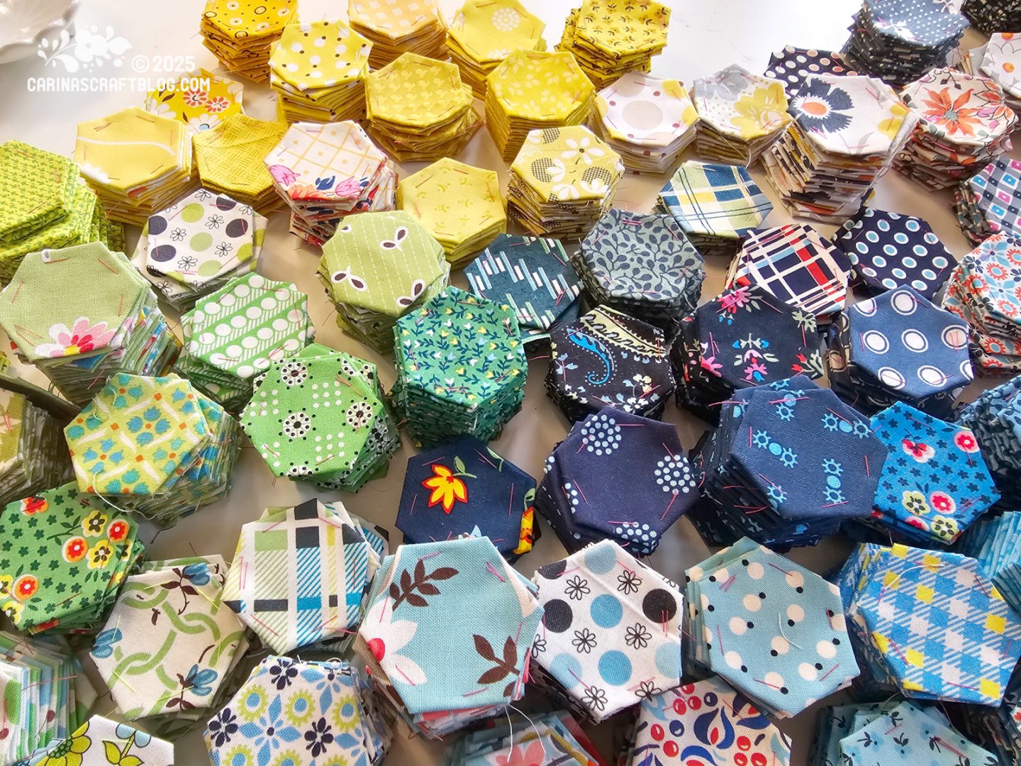 Photo. Close view of a table with stacks of fabric hexagons sorted by colour, mostly yellow, green and blue.