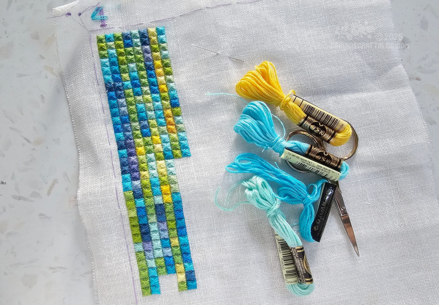 Photo. Close overhead view of a white piece of fabric where columns of square embroidery stitches are worked in different yellow, green and blue colours.