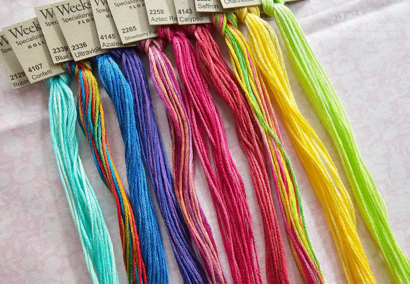 Photo. Overhead view of skeins of colourful embroidery thread laid out next to each other in a sort of colour order from blue to yellow.