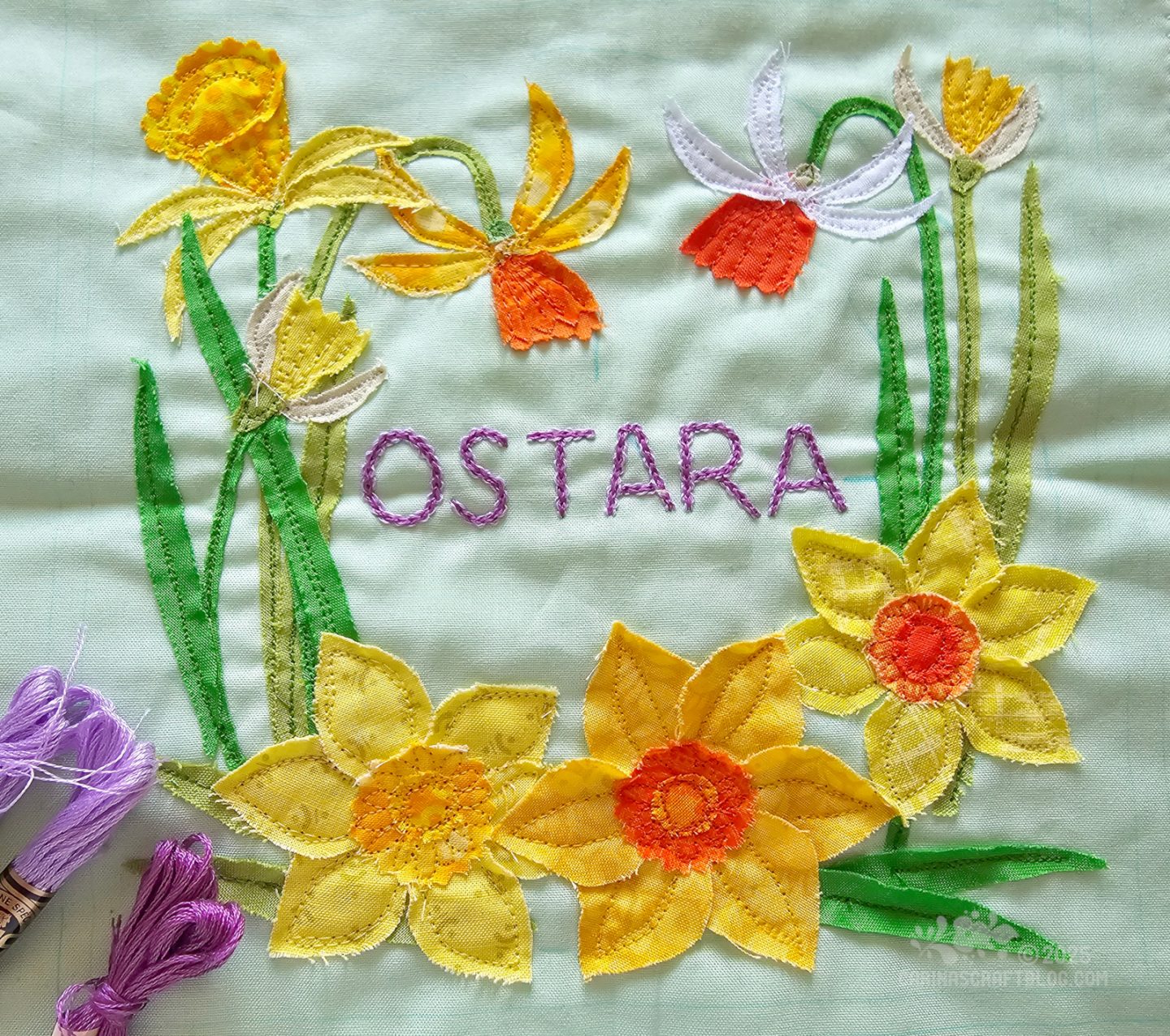 Photo of a square piece of light green fabric. On the fabric are appliquéd daffodils in a sort of circular shape. In the centre the word Ostara is embroidered with purple thread.